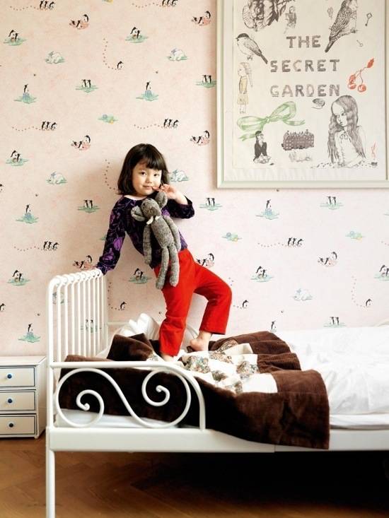 A young girl wearing red pants is posing on a bed with a brown and white blanket in front of a poster of the secret garden.