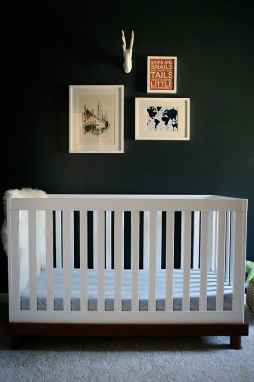 A white crib sits near a mallard green wall that has a few pieces of wall art clustered together on it.