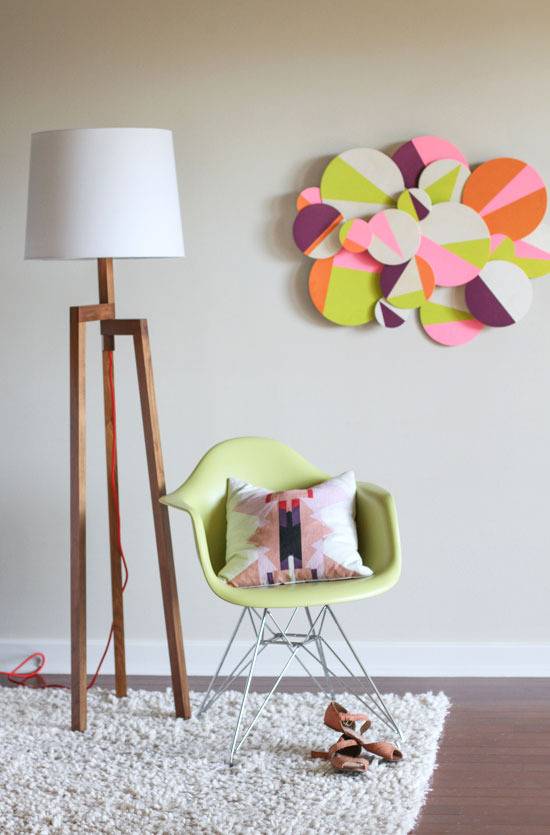 A green chair with a pillow on it, a floor lamp with three wooden legs on a lush white rug, and a green, pink, white and purple circle design hanging on the wall.