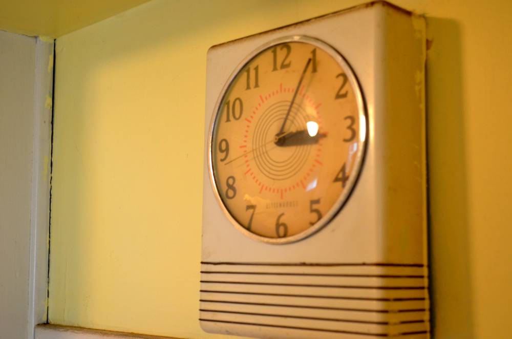 A 20th century wall clock with a speaker hangs on a yellow wall near a ceiling.