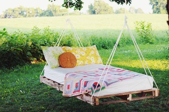 A pallet bed hanging from a tree with flowered pillows, an orange round pillow and a quilt laying over it.