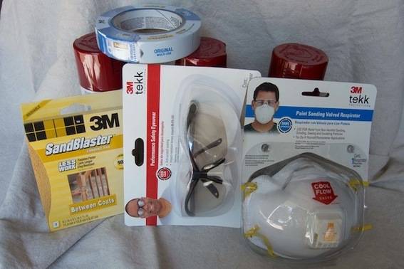 A set of packaged products, a mask, safety glasses, sandpaper, tape and spray paint bottles laying on a white cloth.