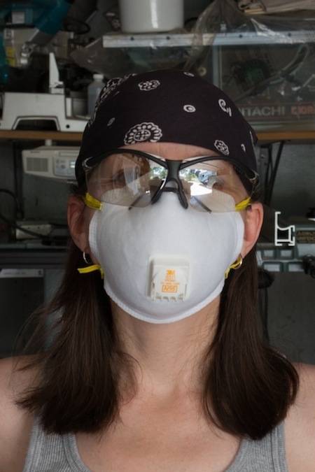 A woman in a gray tank top wears a white mask covering her nose, mouth and cheeks, as well as safety glasses and a black bandana on her head.