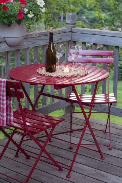 A small round red metal table on a fenced deck, with a wine bottle and two wine glasses on it, and two red chairs around it.