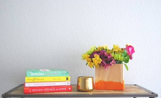 A table top with four books laying sideways, a gold cup and a peach bag with an orange bottom is filled with yellow, purple, green and pink flowers.