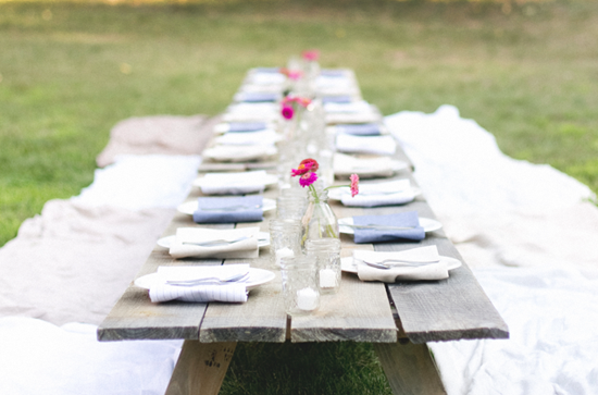 Vases of flowers near folded napkins on long, wood table in the outdoors.