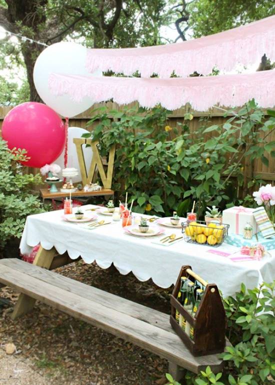 An outdoor picnic table set for a birthday party with pink decorations, a ed baloon and a wooden box of bottles on the seat.