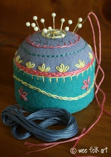 An embroidered pincushion has sewing pins and a sewing needle in it.