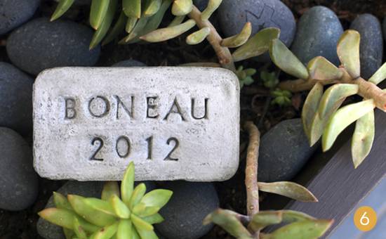 A rectangular stone that says Boneau 2012 on top of some black round rocks and some succulents.