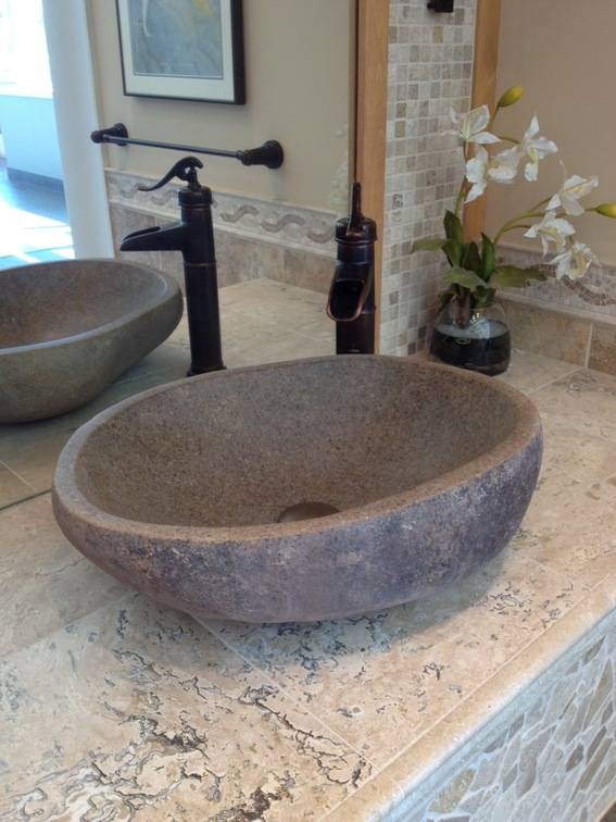 A bathroom sink is seen from an angle and is a light,textured material, with the dull brown clay biwl reflected in the mirror and a tall, black metal faucet with pump handle and a small glass vase with white orchids in a corner.