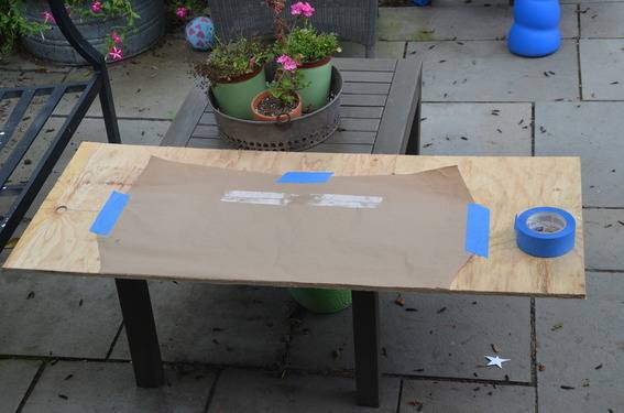 A piece of brown paper taped on wood with blue tape on a patio.
