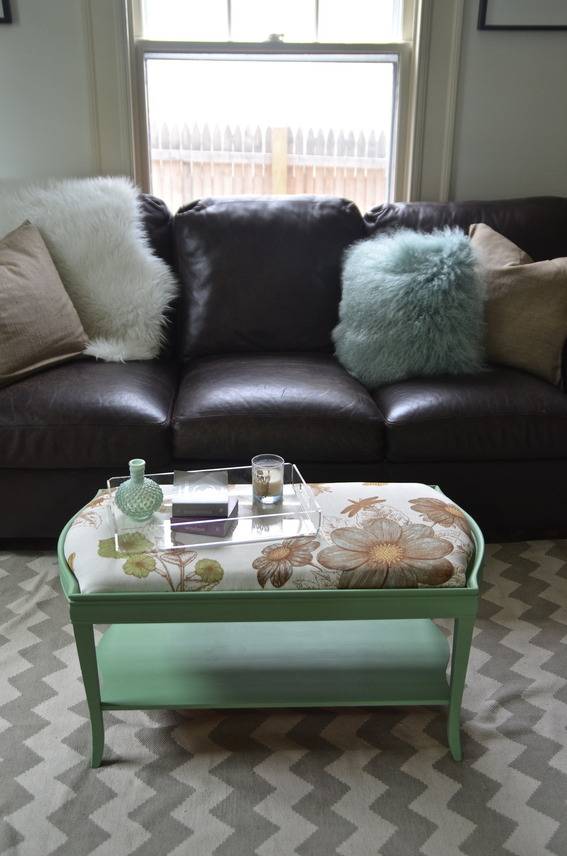 A sofa sits behind a coffee table that has a padded top.