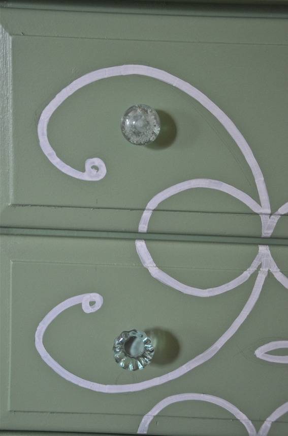 A piece of furniture has been painted green with a white design and has mismatched clear knobs.