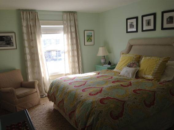 A bedroom with light green walls has a large headboard on a bed and an armchair.