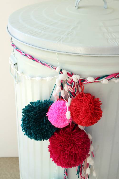 A garbage can painted in white decorated with pom poms.