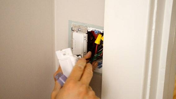 A person is touching the wiring behind a wall switch plate that has been pulled out of the wall.