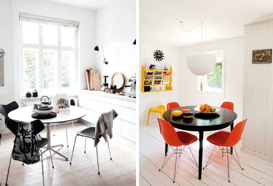 A white kitchen area, with a table in the middle, having both orange and black chairs.