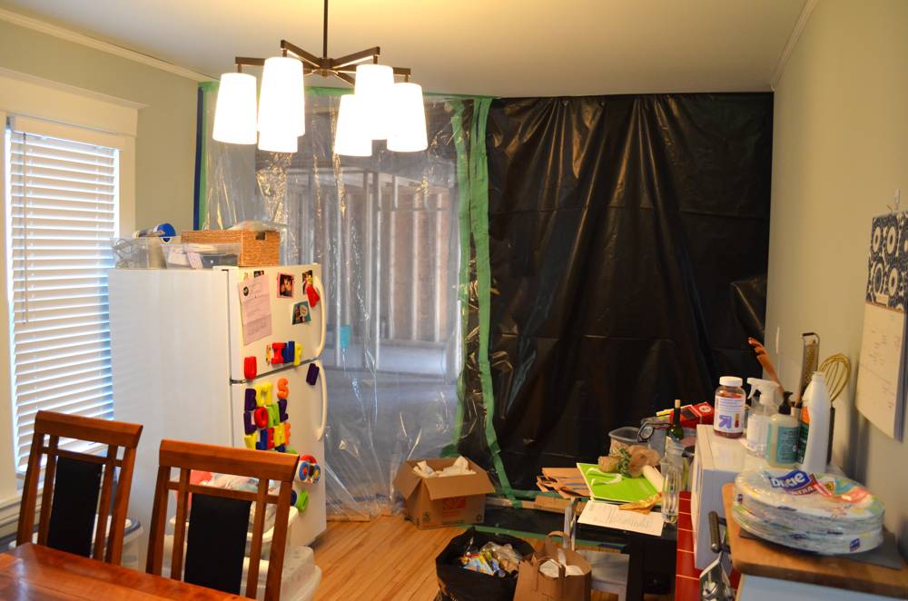 Plastic sheeting covering a wall next to a refrigerator with magnets next to a cluttered counter top in a kitchen.