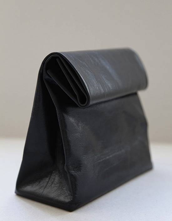 A black leather bag which is shaped like a lunch bag is folded down and seen from an angle on a white table and beige wall behind.