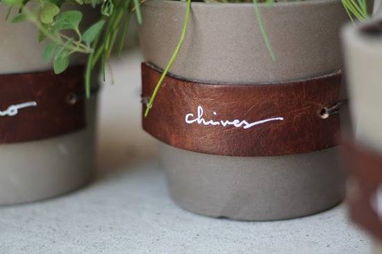A beige flowerpot is seen close up with a brown leather label strapped on that says “chives” in white cursive with chives hanging down;other similar pots are next to it but not in focus and not seen in their entirety.