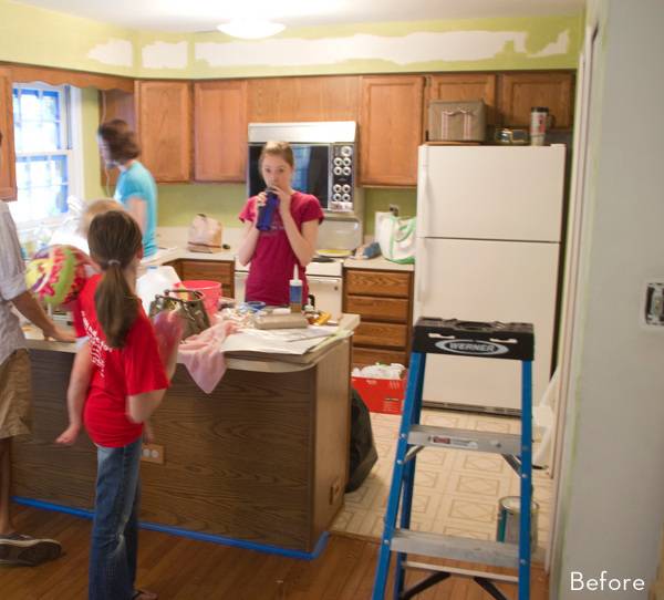 A kitchen with a family in it is taped ready to be painted, there is a blue ladder in the entryway.
