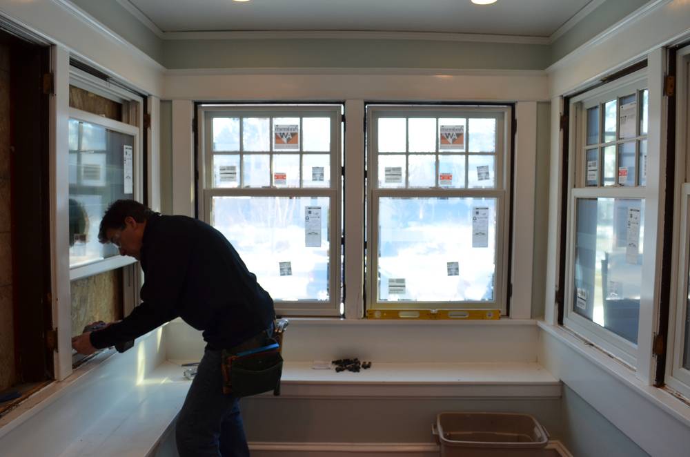 A man fixing a window in a white room with four windows.
