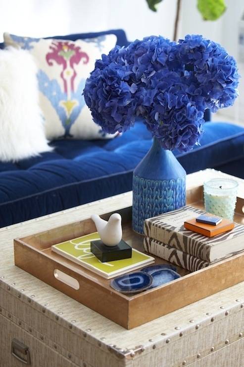 Bouquet of blue flowers in a vase with bird statue and books in a wood tray.
