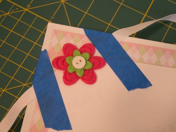 A bordered surface has a ribbon and fabric and buttons flower attached to it with painter's tape.