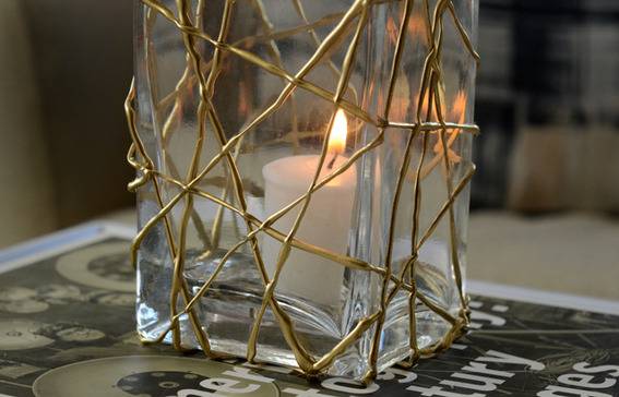 A lit tea candle in a tall rectangular vase with criss crossed gold lines on the outside.