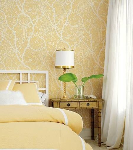 Two large leaves sit in a large vase on a nightstand next to a bed with a yellow bedspread and yellow tree branch wallpaper