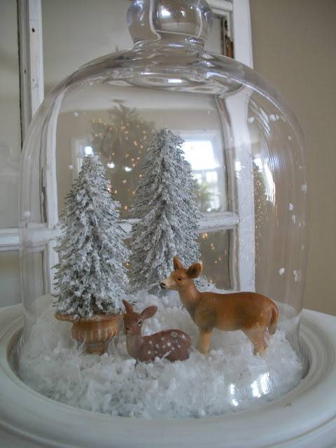 A deer and her fawn standing in deep snow in front of two snow covered trees all under a tall glass dome.