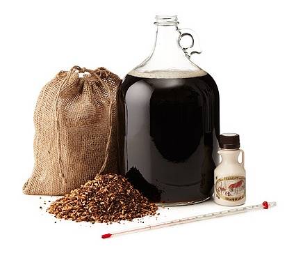Glass growler with dark liquid next to canvas bag and pile of grain.