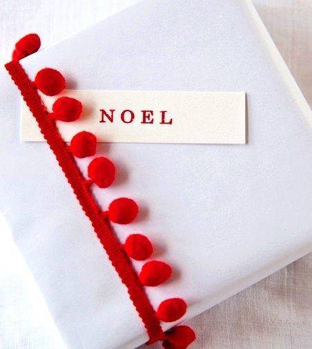 A gift covered in white cloth and a red ribbon with fuzzy red balls and a noel tag slipped under the ribbon.