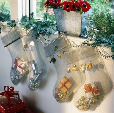 Four transparent stockings hanging from a window sill with gifts in them under a silver box with red flowers.