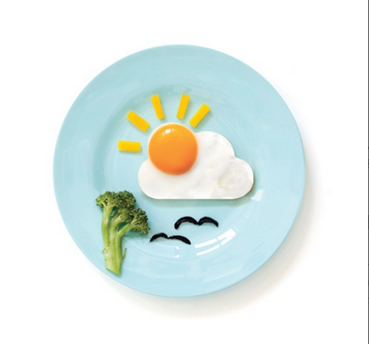 Lines of cheese sit above a sunny-side up egg designed to look like the sun in front of a cloud, and a broccoli below as a tree, with the black olives as birds.