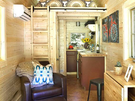 Wooden tiny home with small kitchen and couch, stool, side table with photo and potted plant in hall.