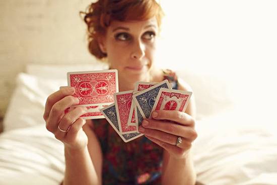 By sitting on the bed, a woman is holding playing cards and looking somewhere else.