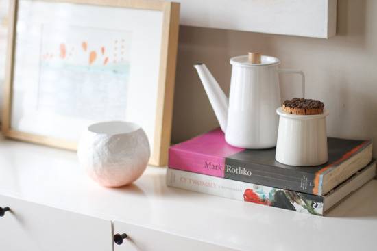 Two books, one kettle, one vase and a pot lying on a shelf near a painting.
