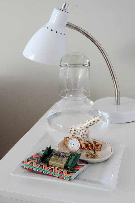 Desk lamp, and side table items like chocolate, jewelry, and cup of water, on a night stand.
