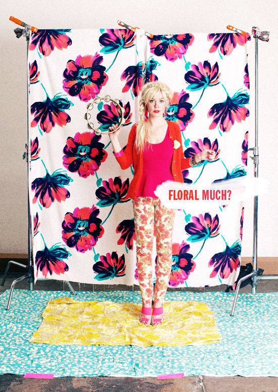 A woman wearing red top and floral pants is standing in a room where floral sheet is on her bottom and back as well.