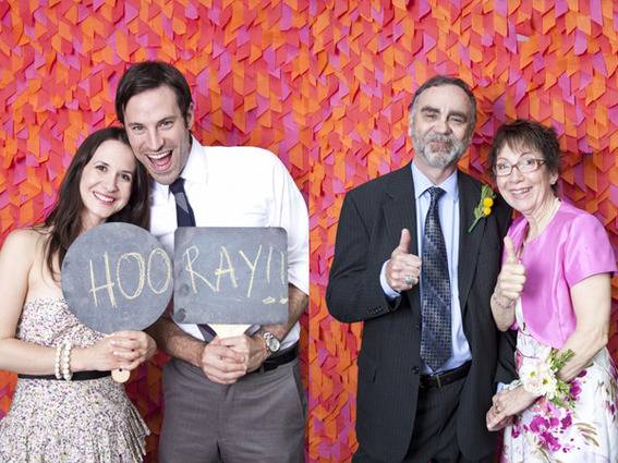 A man and woman hold up a sign that says hooray in front of a confetti backdrop, next to an older couple holding up thumbs