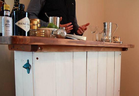 A wooden bar with bottles of alcohol, wine glasses, a garlic press and a man's torso.