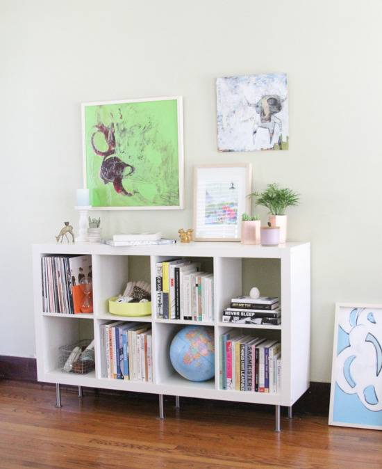 White color bookshelf with lots of books and potted plant at top and painting frames on wall.