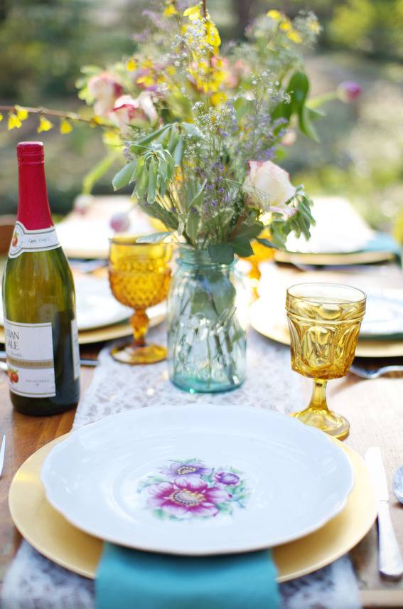 A table with wine, plates and a vase full of flowers on it.