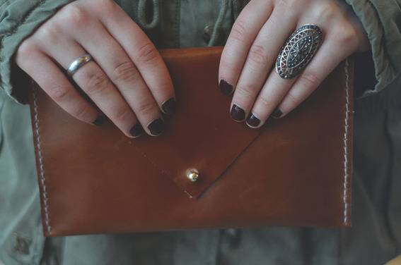 Two hands with black nail polish are holding a brown clutch purse.