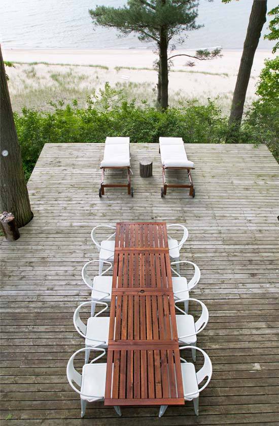 A brown slat table with eight white chairs in front of two lounge chairs on a deck overlooking a beach.