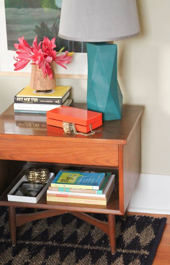 A wooden night table that has books, a jewelry box, and a blue angular lamp.