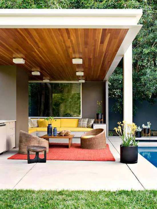This modern patio features a wood ceiling with lighting, a large red area rug, yellow sofa, and simple wicker-looking chairs.