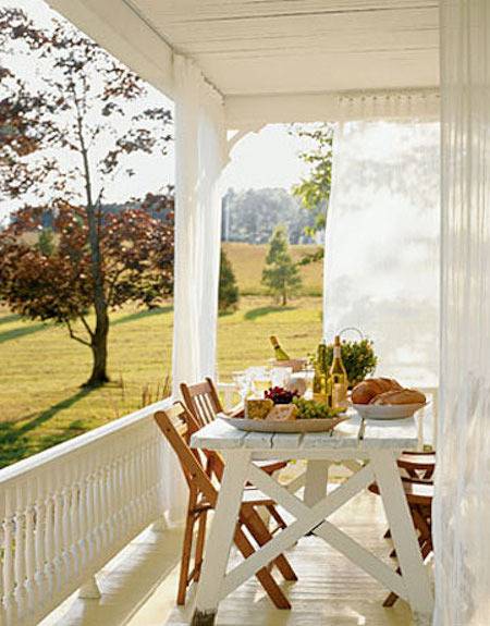 A white porch on a sunny day with sheer curtains and a white table with brown chairs set with bread and cheeses.