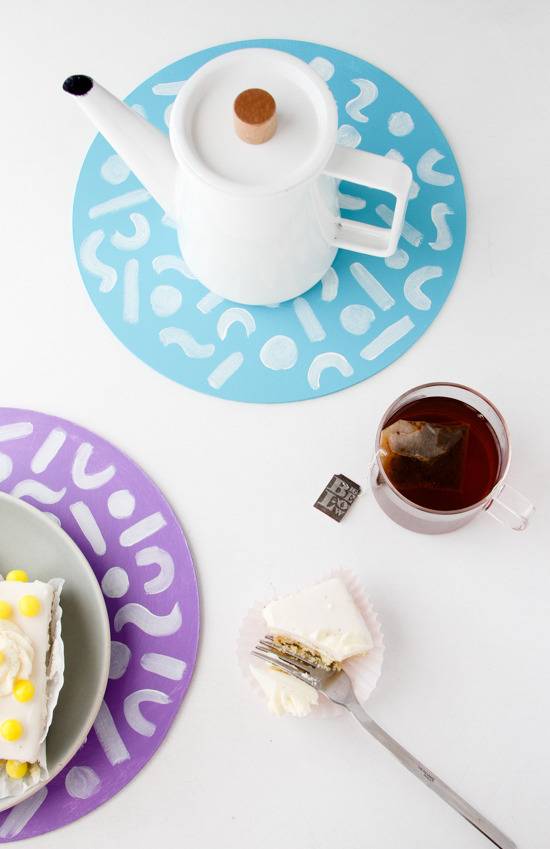 A white coffee warmer on a blue circle with white designs, a cup of tea, a fork left in a petite four and part of a dish on a purple circle with white designs.